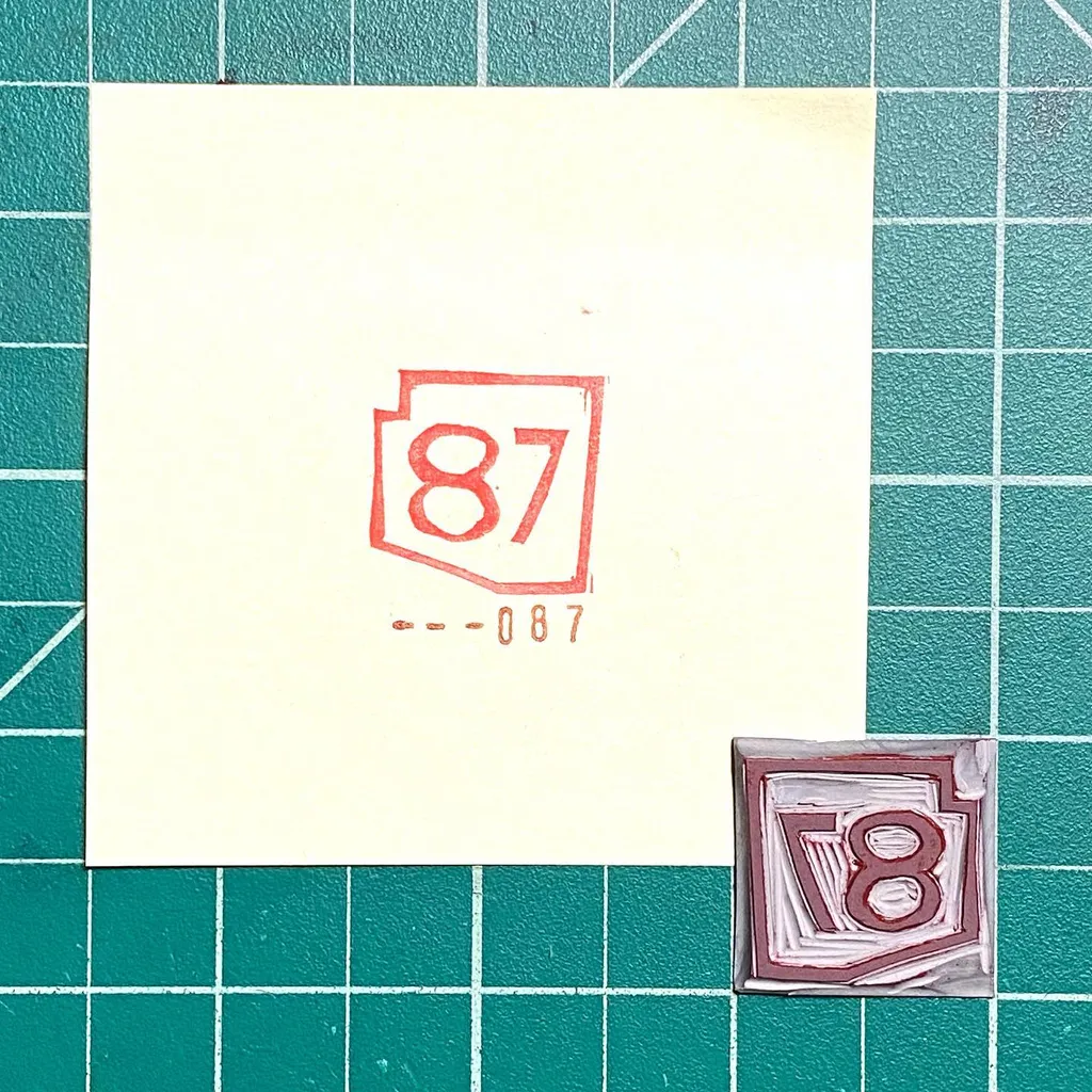 A cream paper on a teal grid displays red numbers 87 in a box, with --087 written beneath it, next to a small, painted square featuring the number 8 in reddish-brown tones.