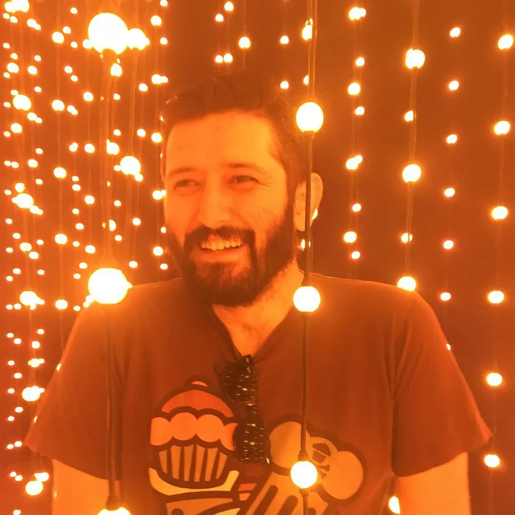 A smiling man with a beard wears a dark t-shirt against a background of dense, glowing warm yellow and orange string lights.