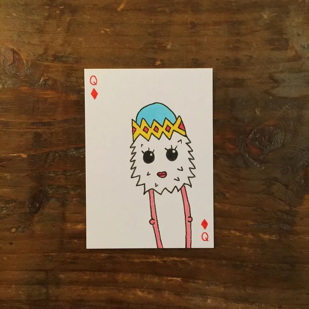 A cartoon queen drawing on a playing card, featuring a light blue crown and a jagged white wig, with pink elements hanging below, set against a dark wooden background.