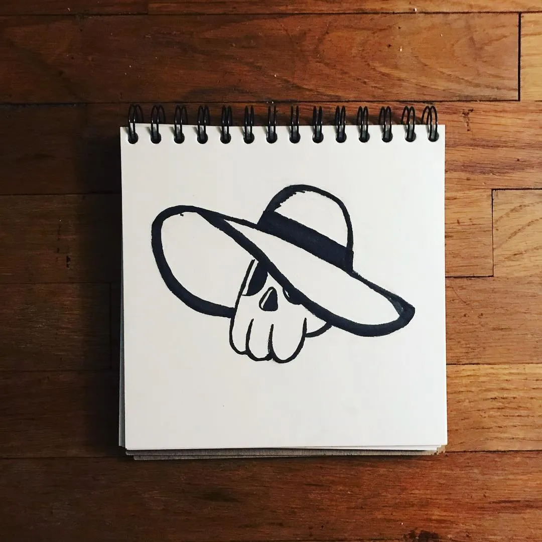 A black line drawing of a skull wearing a wide-brimmed hat is centered on a spiral-bound sketchbook resting on a wooden surface.