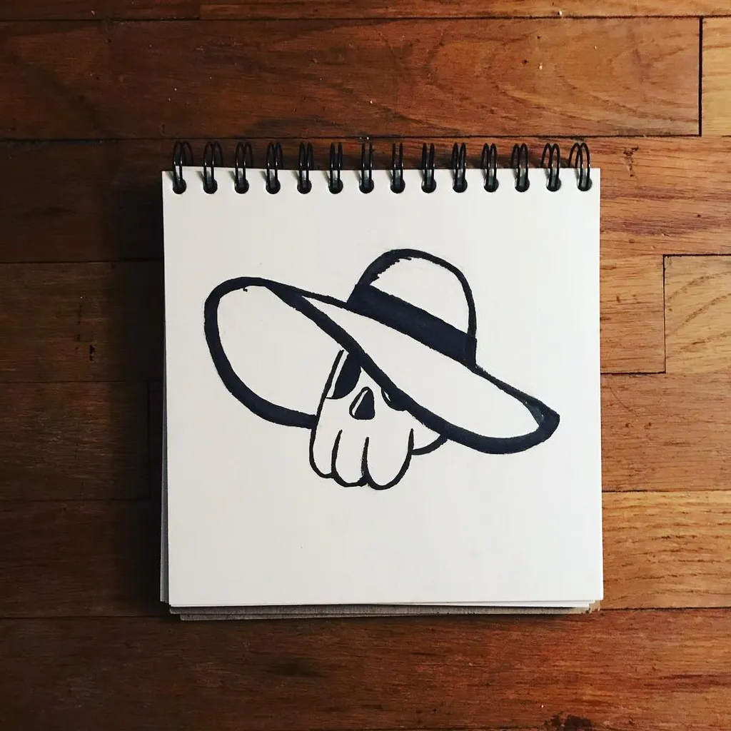 A black line drawing of a skull wearing a wide-brimmed hat is centered on a spiral-bound sketchbook resting on a wooden surface.