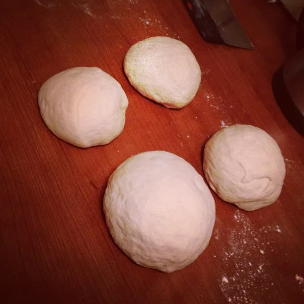 Five pale, smooth mounds of dough are arranged on a reddish-brown wooden surface dusted with white powder, with a dark metallic object visible in the upper right corner.