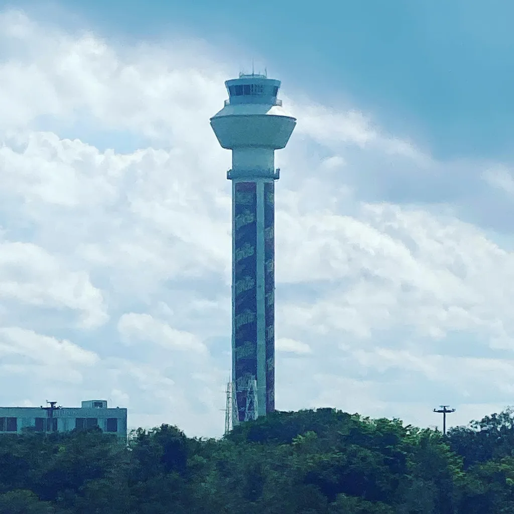 A tall, patterned tower rises from dense dark green trees against a light blue, cloudy sky, with smaller structures visible on the left.