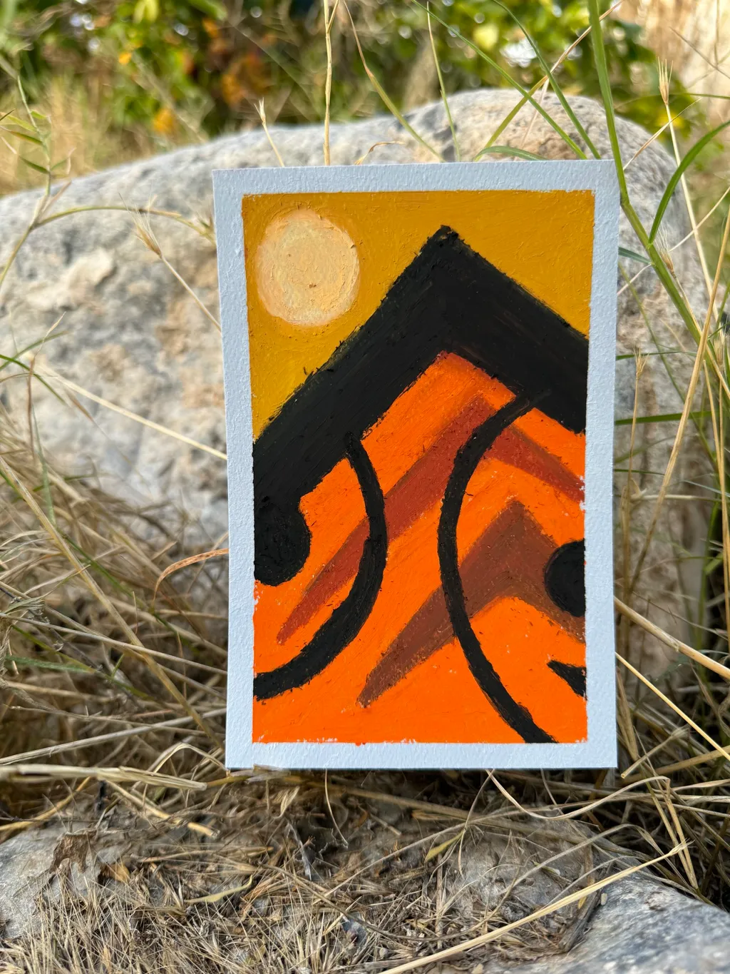 A small, vertical artwork featuring abstract orange and black geometric shapes, topped by a black triangle and accented by a yellow circle, rests on rough gray rocks and dry grass.
