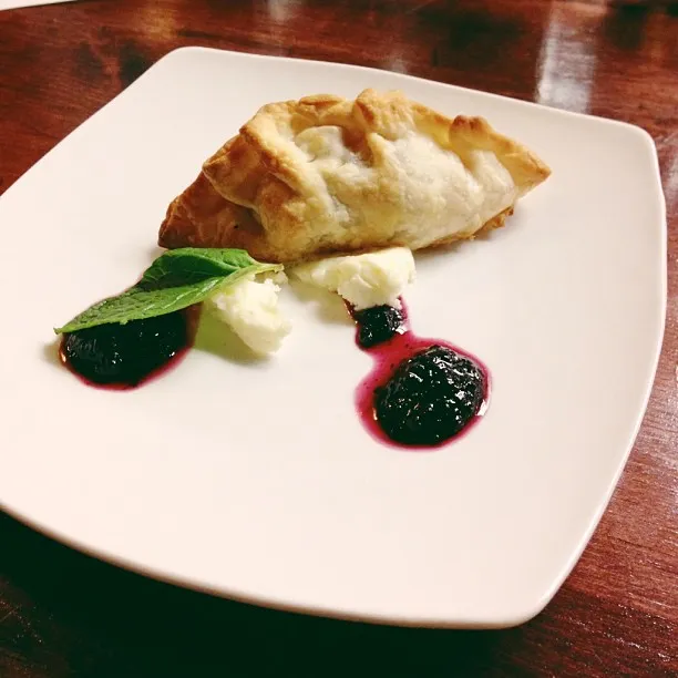 A square white plate displays a golden-brown, flaky pastry piece arranged above three distinct groupings of dark purple sauce, white cream, and a green leaf.
