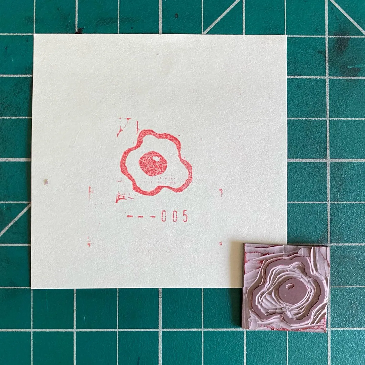 On a teal grid, an off-white card displays a red outline of a lobed flower and the number --005, positioned next to a small, textured pink square featuring concentric, layered patterns.