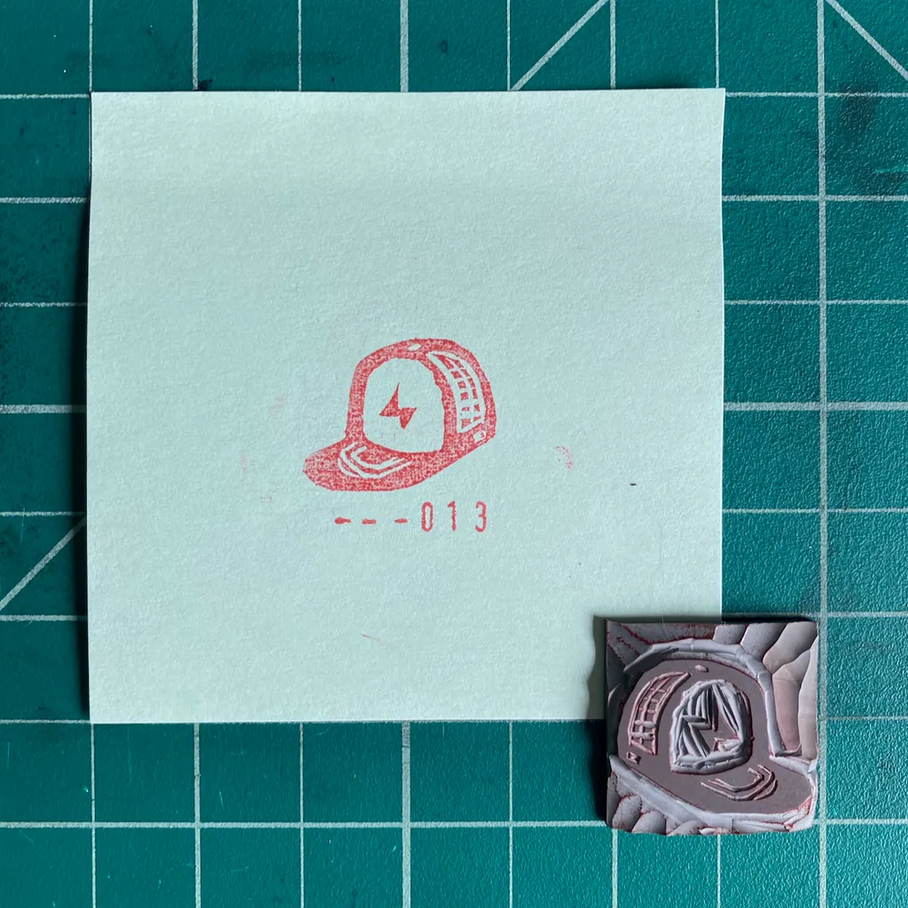 A cream card on a dark teal grid shows a red drawing of a baseball cap with a lightning bolt and the numbers --013, next to a small, molded reddish-brown cap object.