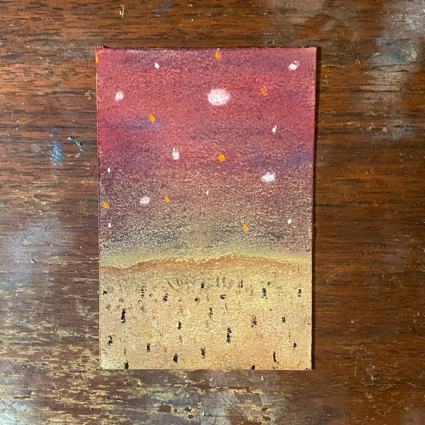 A rectangular artwork depicts a layered scene, transitioning from a speckled reddish-maroon sky with white spots, through a gradient band, to a tan foreground marked with dark vertical lines.