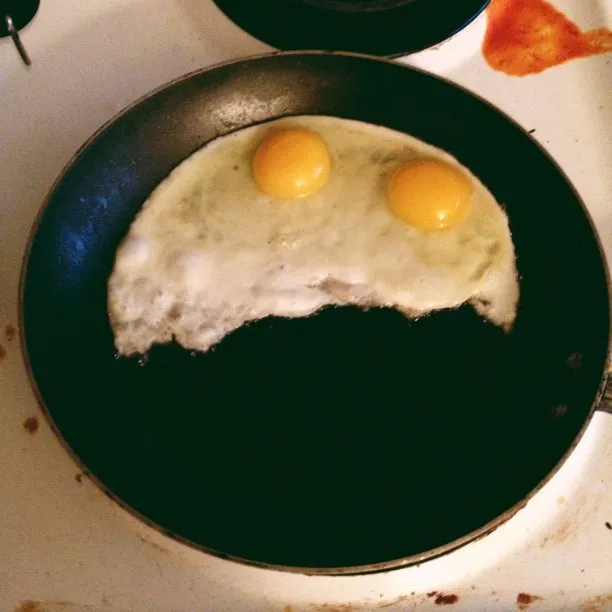 Two pale, round fried eggs with bright yellow yolks are centered in a dark, circular pan resting on a white stove surface marked with reddish-brown spatters.