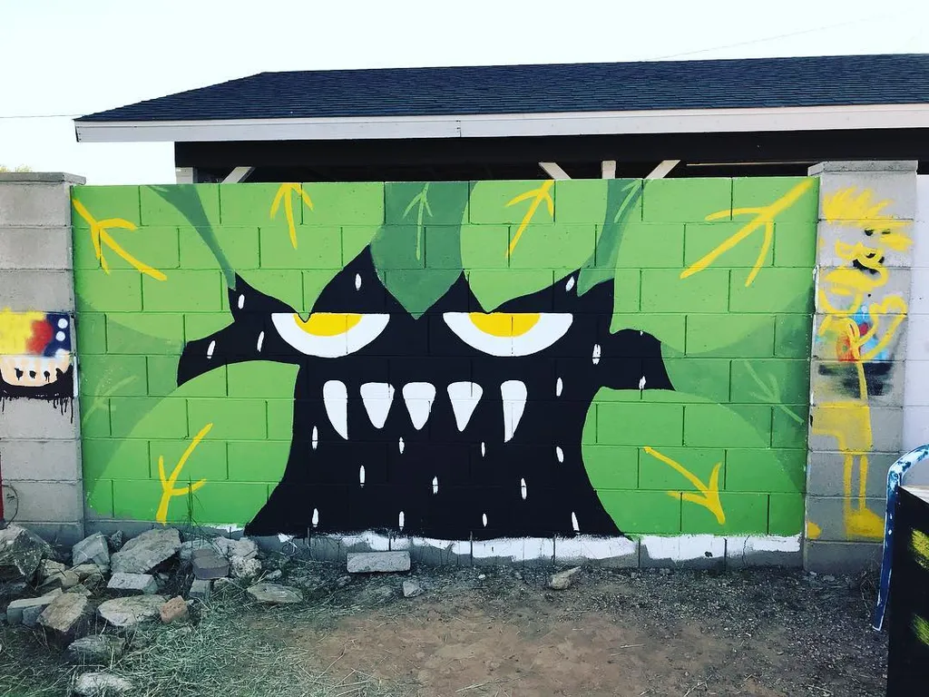 A large mural features a stylized black monster face with three yellow eyes and white teeth, painted on a bright green background patterned with yellow lines.