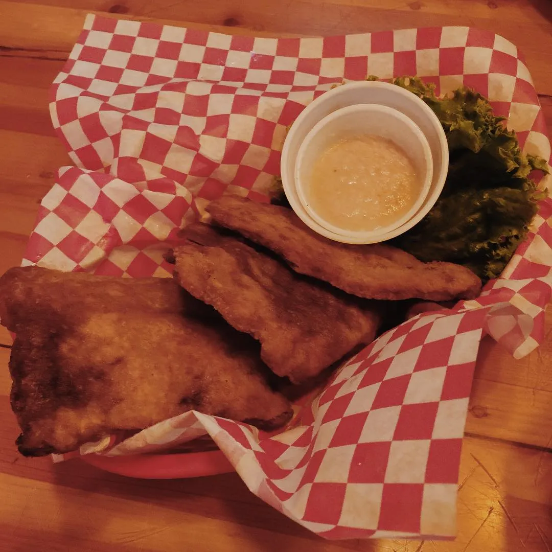 Several golden-brown, fried pieces rest on a red and white checkered liner on wood, next to a small cup of light yellow sauce and dark green relish.