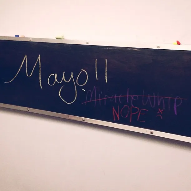 A dark rectangular surface mounted on a light wall displays handwritten marks in white and pink, forming text such as Mayo 11 and NOPE.