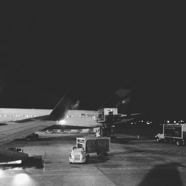 Dark night scene on an airport tarmac featuring several parked service vehicles and a distant runway, illuminated by pools of yellow and white artificial light.