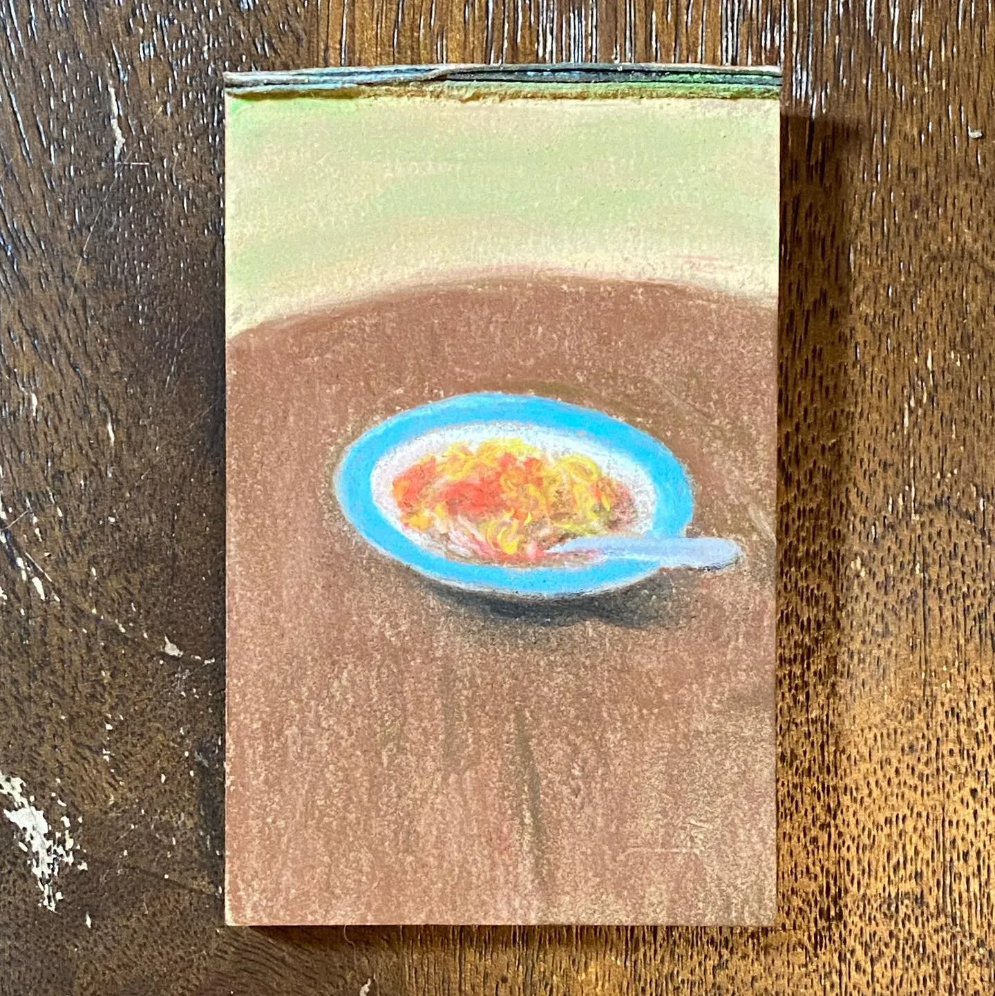 A vertical painting features a blue-rimmed bowl holding yellow and orange food, with a spoon resting nearby, set on a brown surface beneath a gradient of light green and tan.