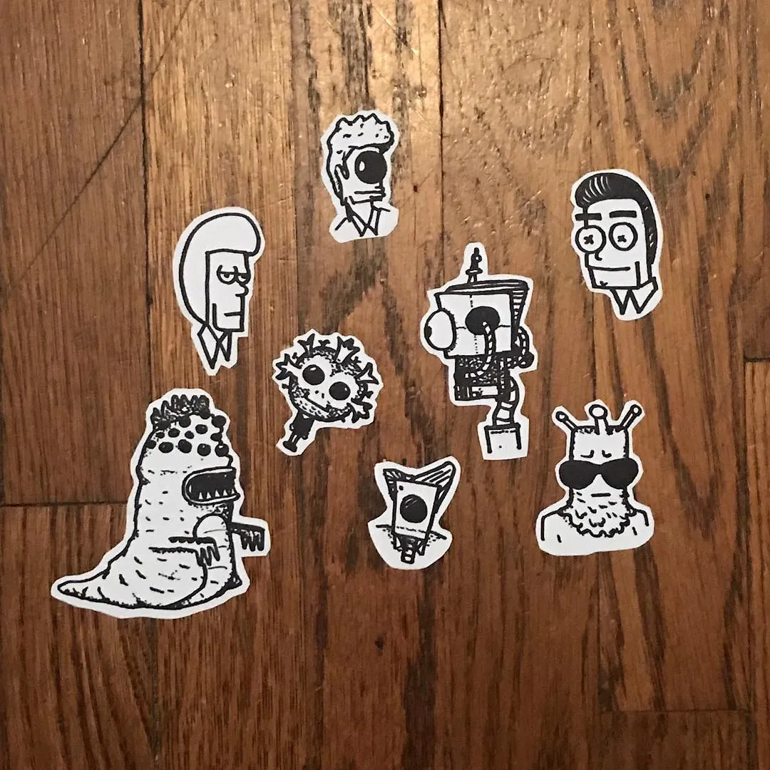 Multiple black and white line drawings of diverse cartoon characters, ranging from a dinosaur to a robot, are arranged randomly on a wooden surface.