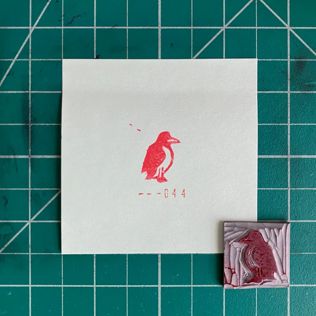 A white card on a teal grid displays a red stylized bird and the numbers --044, positioned next to a small, textured square of layered red and pink material.