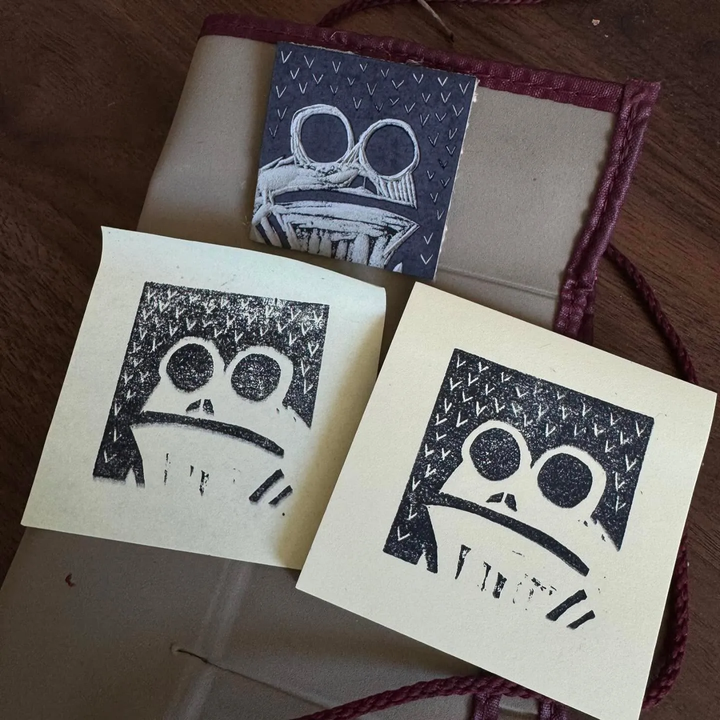 Three small, pale cards featuring black ink drawings of stylized faces are arranged on a closed journal with a reddish cord, set against a wooden background.