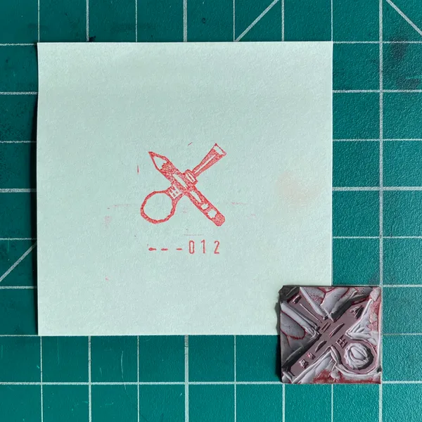 A light mint square paper on a teal grid background displays a red drawing of crossed tools and the numbers --012, positioned above a small reddish-brown square object with an embossed mechanical design.