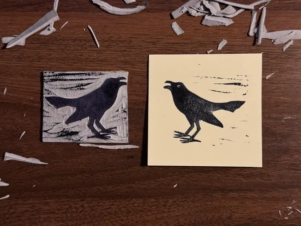 Two black bird silhouettes are displayed on a wooden surface, each on a square piece of paper; the left background has distressed horizontal lines, and the right background has faint horizontal marks.