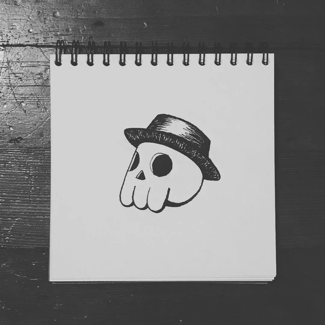A black and white drawing of a stylized skull wearing a hat, centered on a spiral notepad placed on a dark, textured wooden surface.
