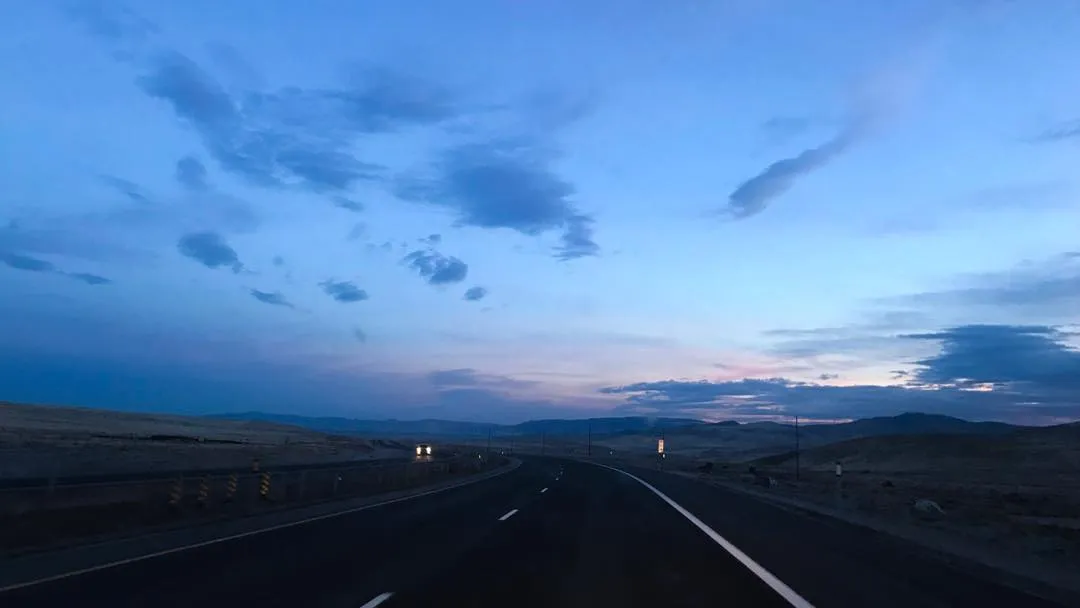 A straight asphalt road leads through a desert landscape under a vast sky of deep blue and gray clouds, with soft pink and orange hues near the distant horizon.