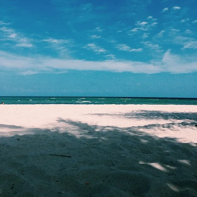 A wide beach scene features a bright blue sky with scattered clouds above a turquoise ocean, meeting a sandy foreground covered in deep, dark shadows.