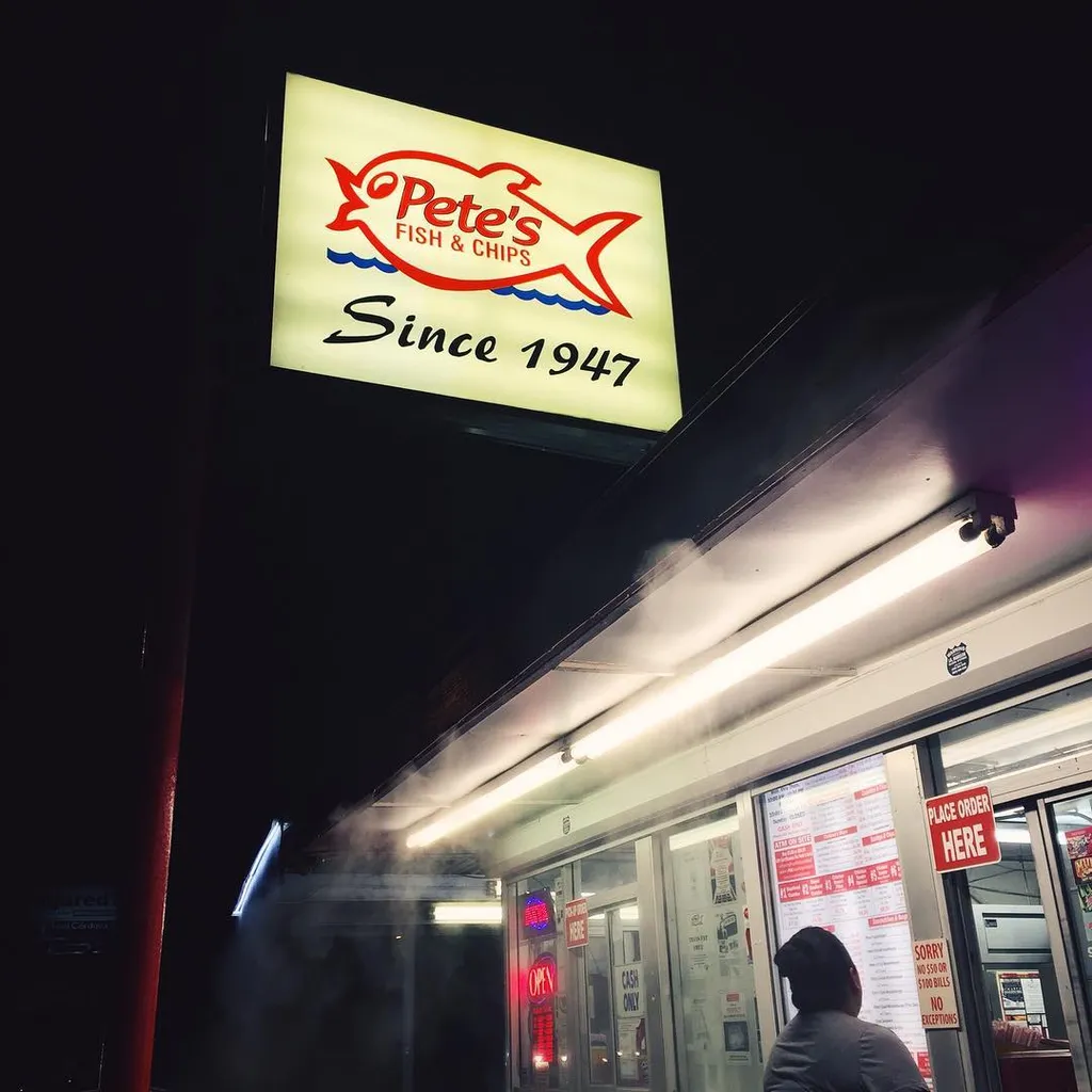 A brightly lit yellow and red sign reading Pete's Fish & Chips Since 1947 hangs from a dark overhang above a shop entrance filled with visible steam and glass windows.