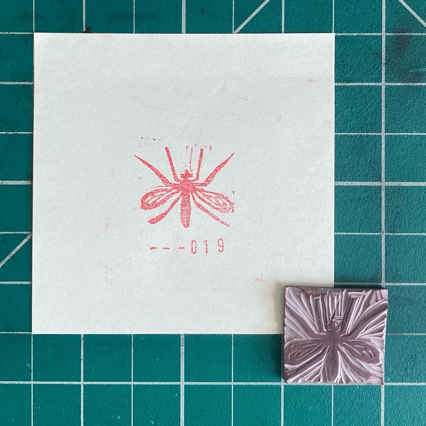 A white square on a teal grid displays a red stylized dragonfly drawing with numbers 610-019, positioned above a small, embossed silver square featuring a detailed dragonfly design.