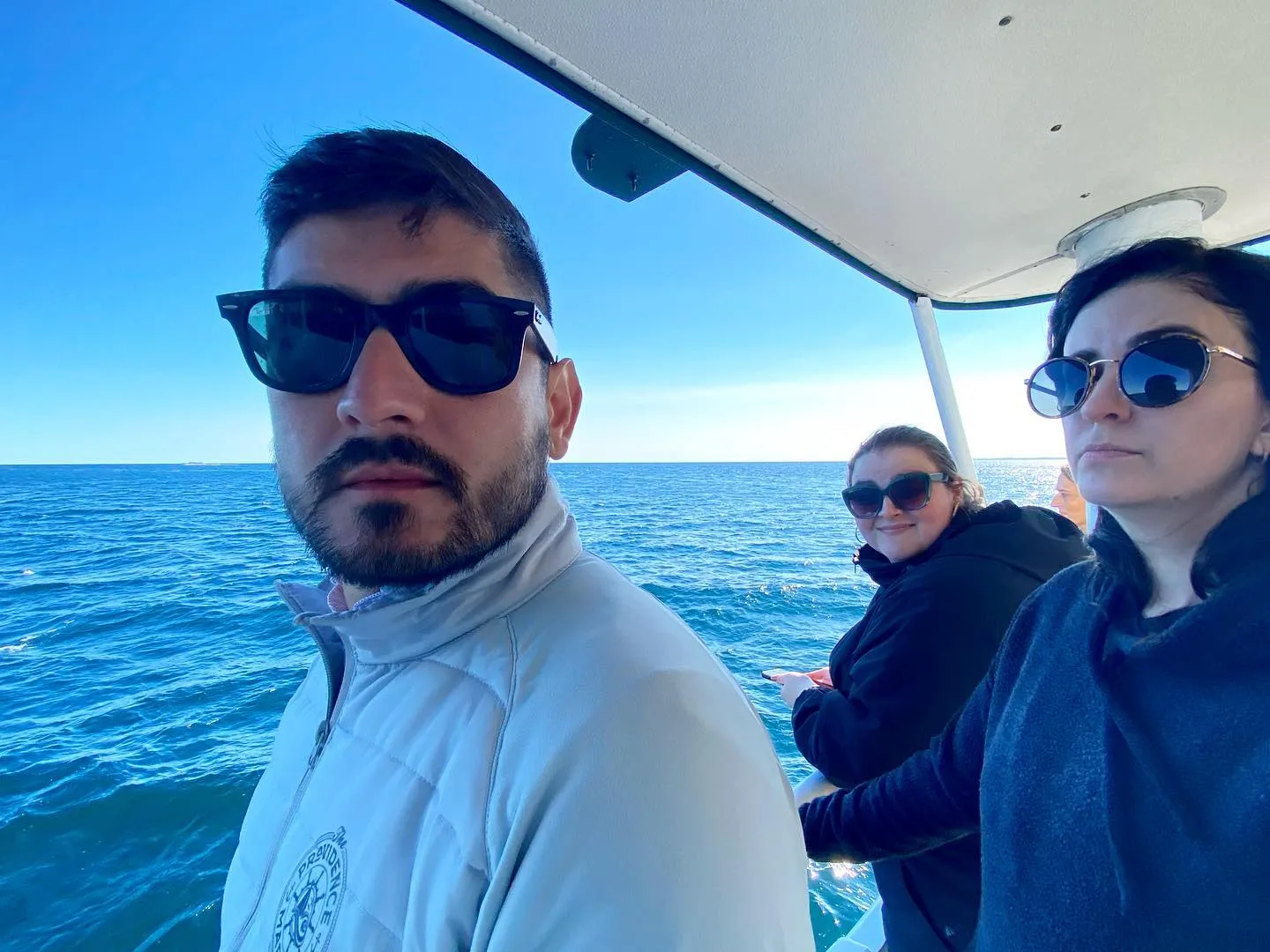 A man in a light blue jacket occupies the foreground, looking out over a vast blue ocean and sky, with three other people visible under the white canopy of the boat.