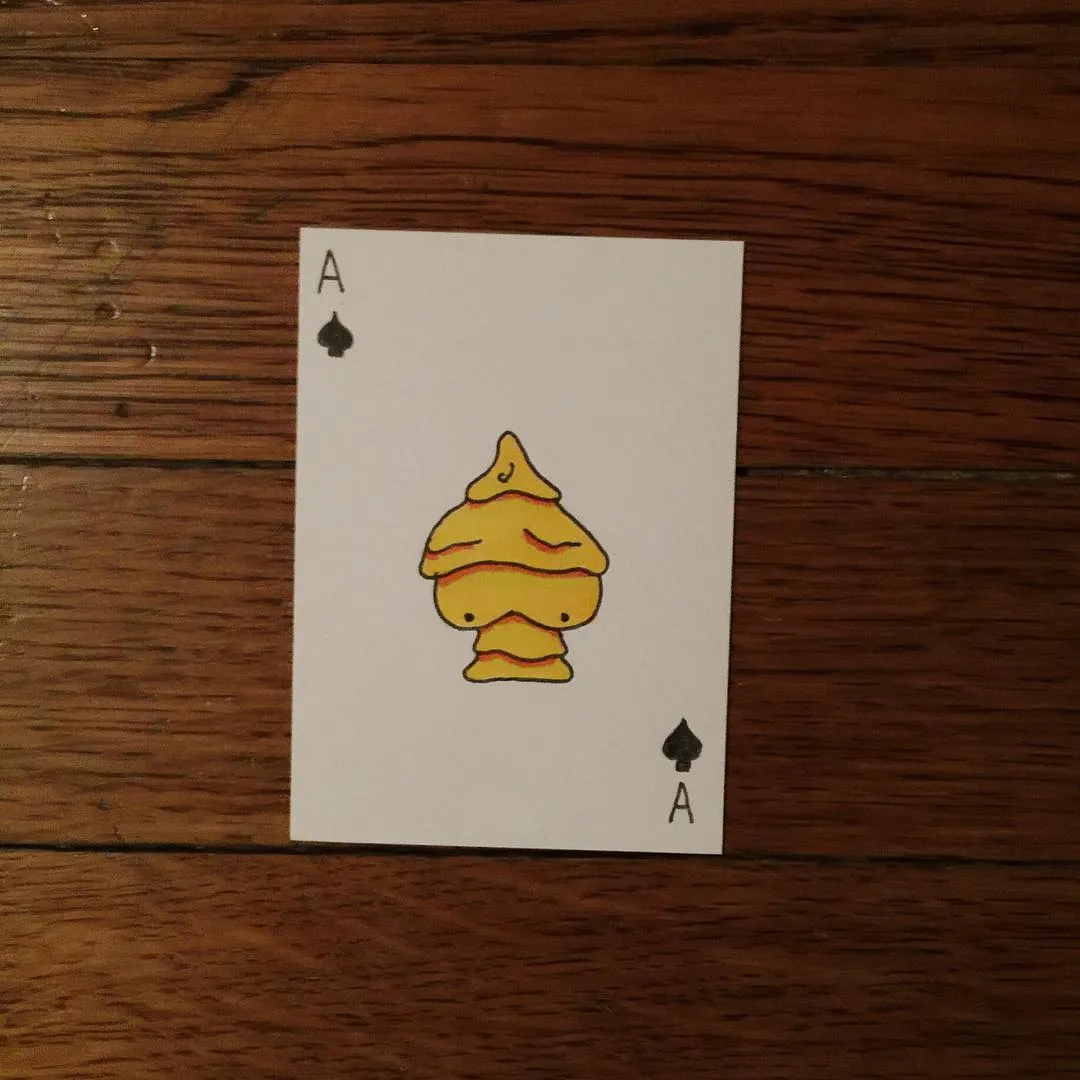 A playing card featuring an Ace of Spades is centered on a dark wood grain surface, displaying a yellow, layered, cartoon figure in the middle.