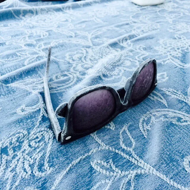 Dark sunglasses lie on a cool blue fabric featuring a repeating, woven pattern of lighter blue lines and soft folds.