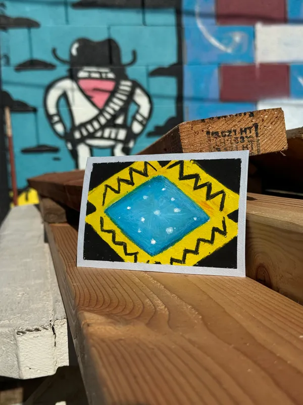 A small square painting with a yellow zigzag border surrounds a blue diamond containing white stars, resting on rough brown wood planks in front of a blurred background featuring a stylized figure.