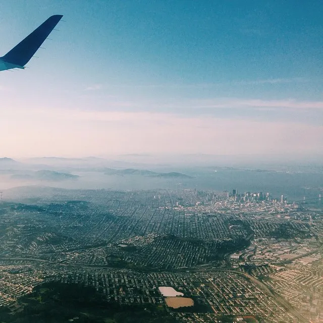 A partial wing occupies the upper left corner, contrasting a bright blue sky with a sprawling, patterned urban landscape that fades into hazy blue mountains on the horizon.