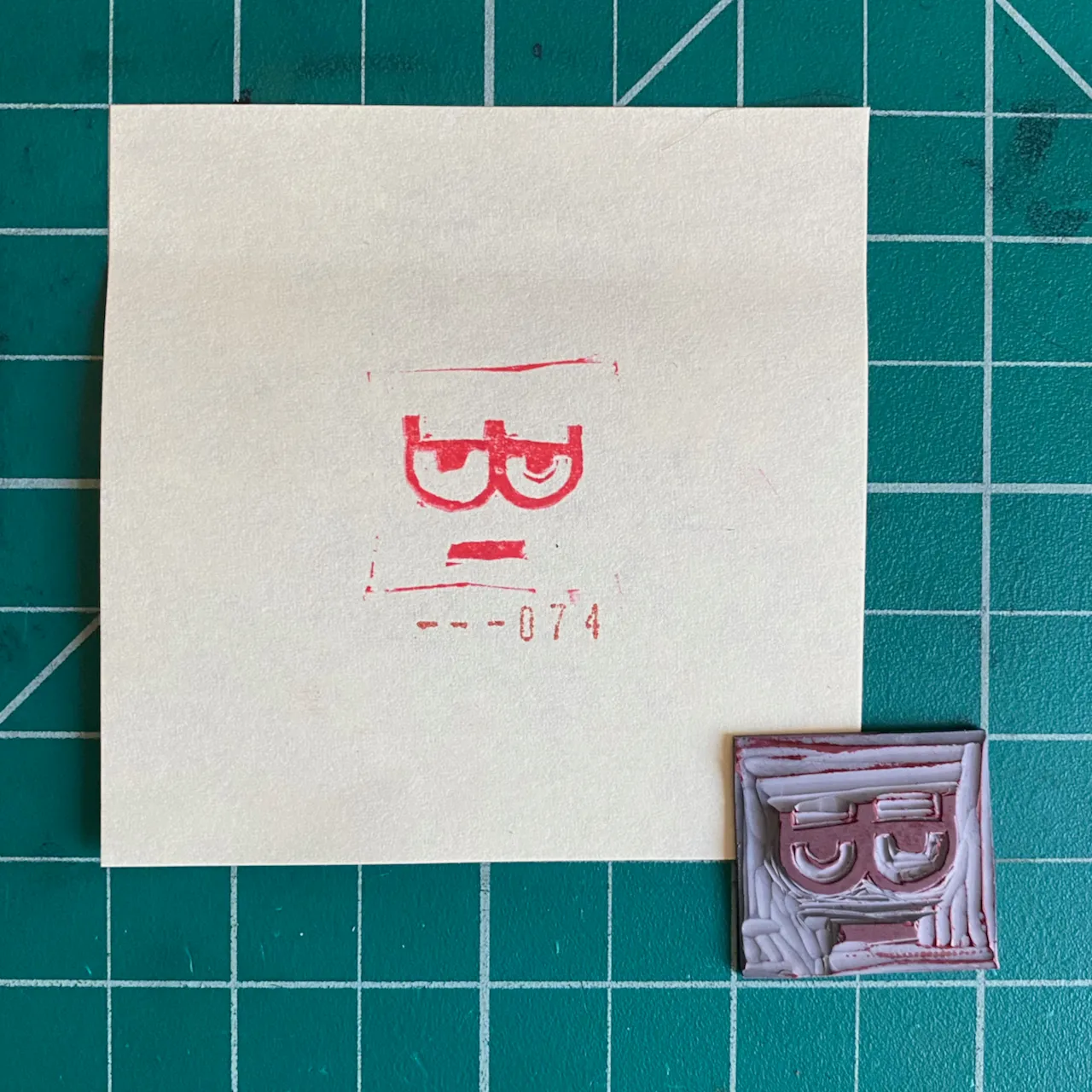 A cream paper rests on a teal grid background, featuring red ink drawings of a stylized face and the number --074, next to a small, rectangular, purple and white molded object with a stylized face.