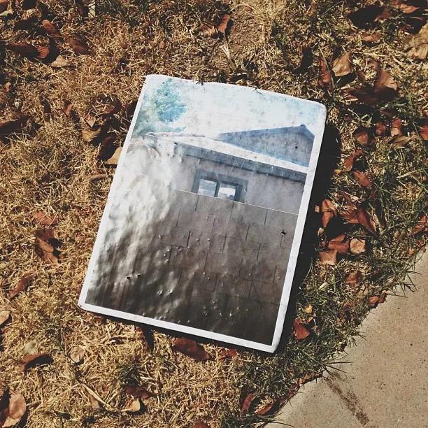 A rectangular, faded photograph depicting a weathered gray wall and a light blue roof rests on a surface covered in dry brown leaves and patchy green grass.