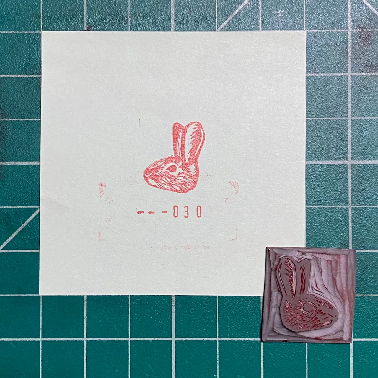 A white paper displays a reddish-pink ink drawing of a rabbit and the number 030, positioned next to a small, reddish-brown carved rabbit object, all set against a teal grid background.