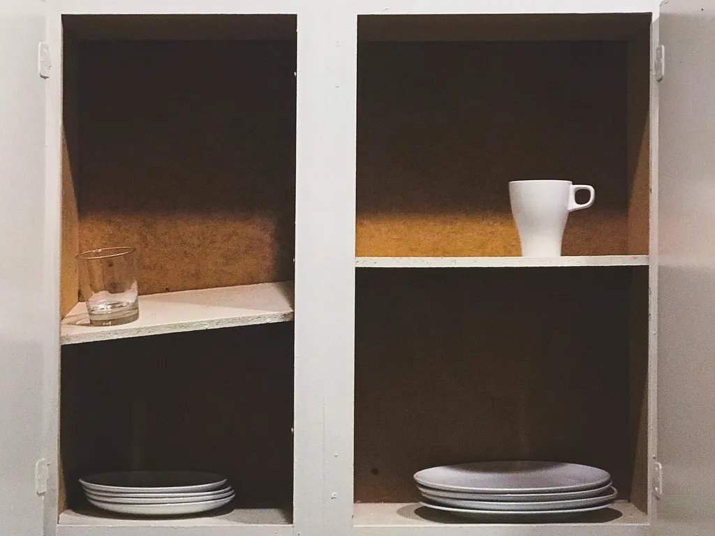 Two white shelving units hold various objects against tan back panels, including a clear glass, a white mug, and stacks of light-colored plates.