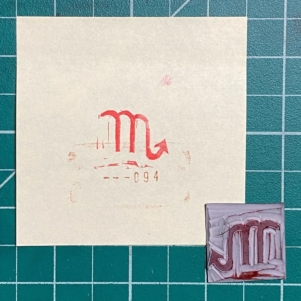 A cream square paper on a teal grid displays a red stylized letter M and the numbers -094, positioned above a small reddish square containing an impressed, blocky letter M.