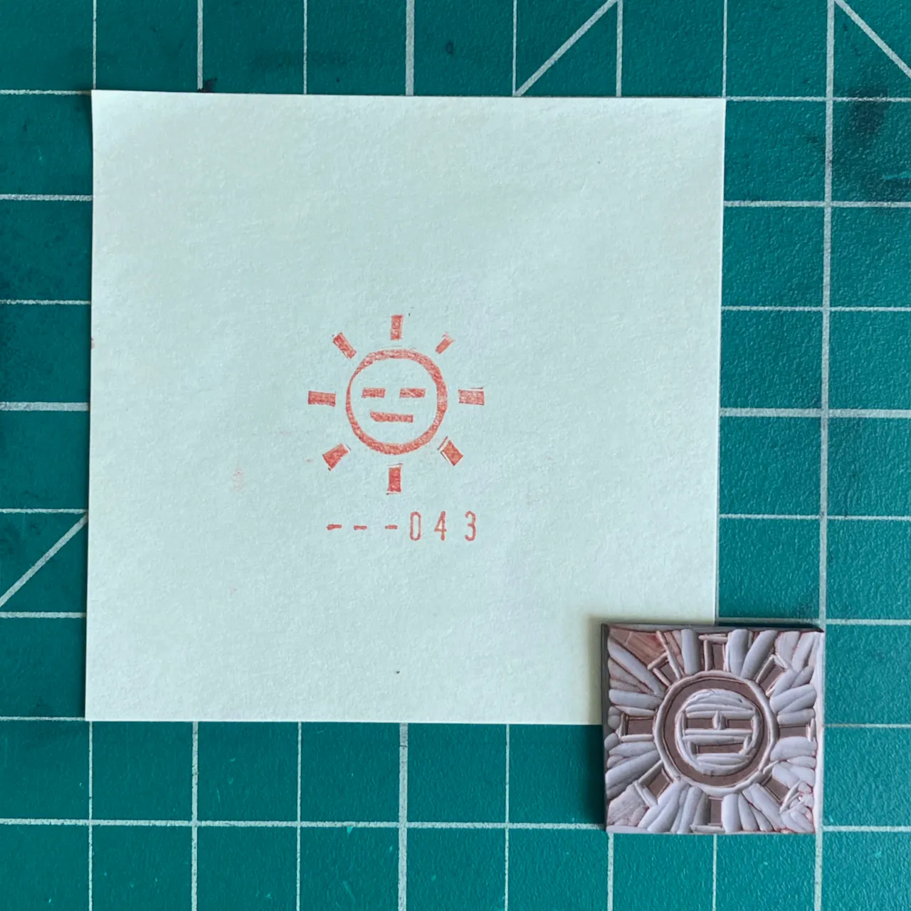On a green grid, a white paper displays a red sun drawing and the number --043, next to a small, textured, light brown square featuring a central circular motif.