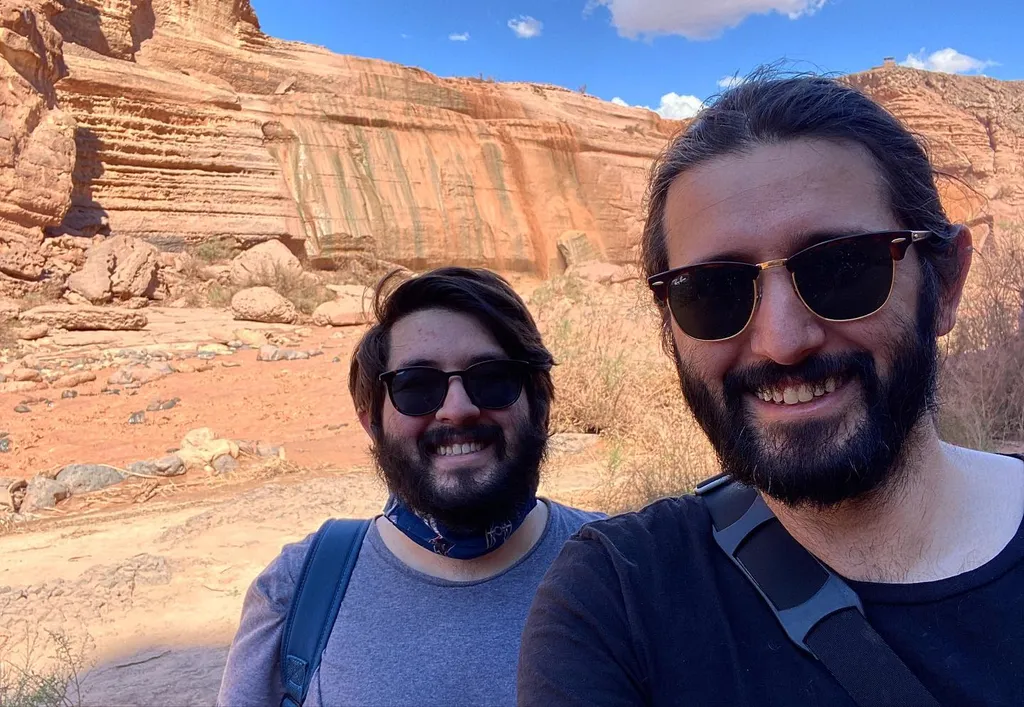 Two men with dark hair and sunglasses smile in the foreground, set against a dramatic backdrop of striated, reddish-orange canyon walls and a blue sky.