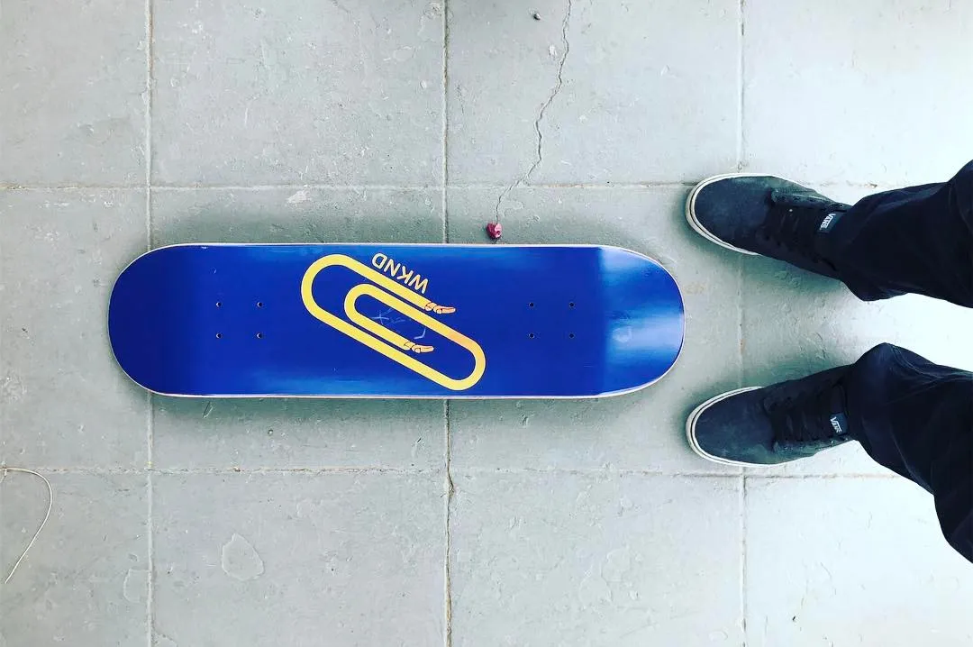 A bright blue skateboard with a yellow graphic rests on a light gray tiled floor, flanked by two pairs of dark shoes.