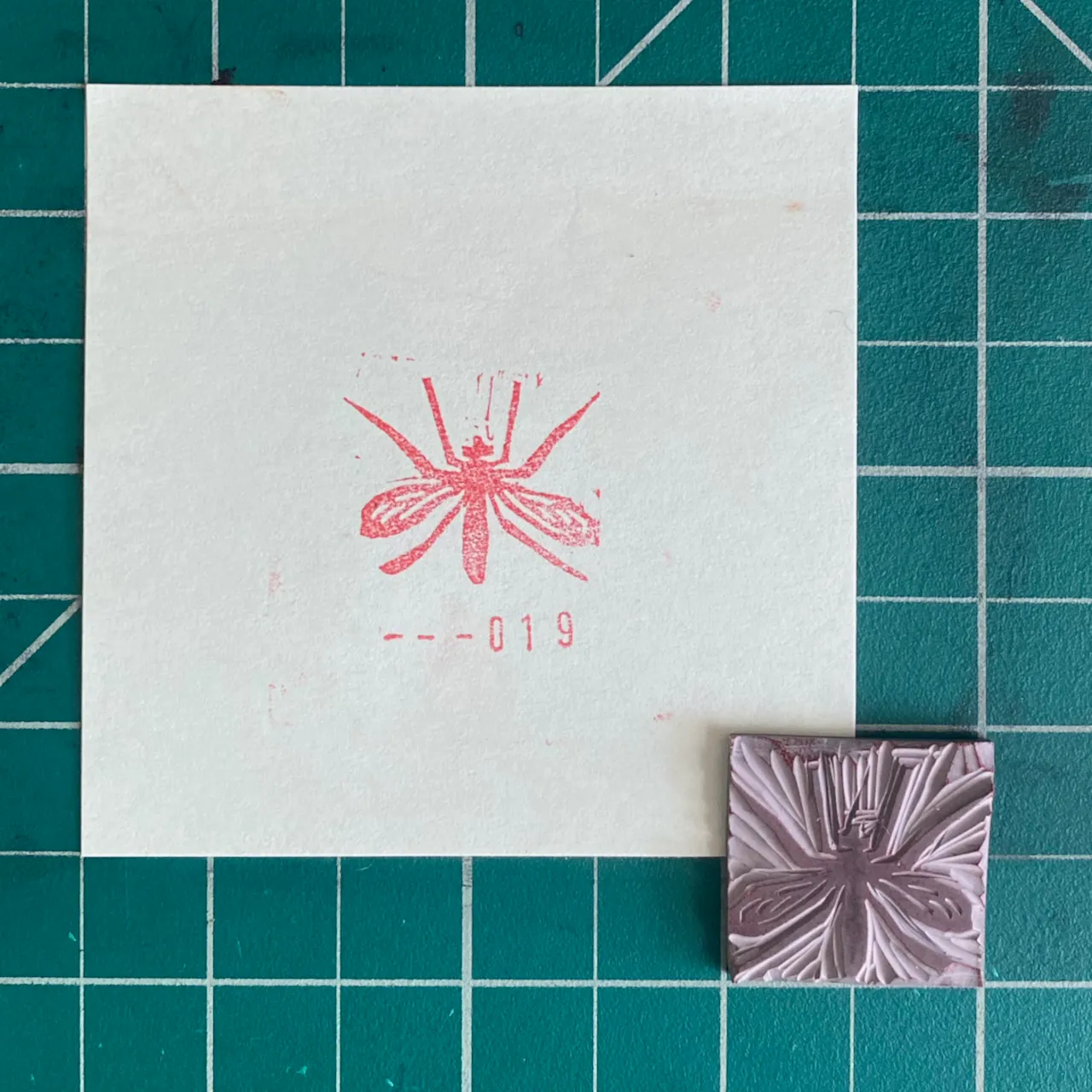 A white square on a teal grid background displays a red line drawing of a spider and the number -019, alongside a small, square, textured purple object in the bottom right corner.