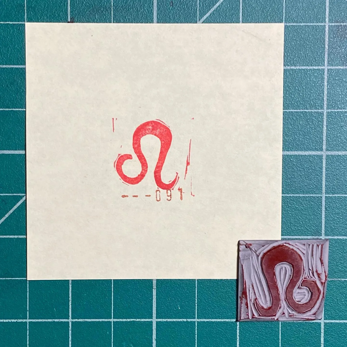 A white square on a teal grid displays a red, looping symbol and numbers, with two smaller, reddish-brown molded pieces featuring similar symbols arranged below.