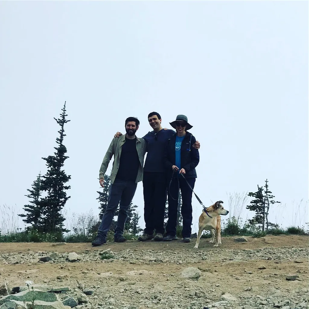 Three men stand on dry, gravelly ground with a dog on a leash; they are positioned beneath a pale, overcast sky and sparse, dark green trees.