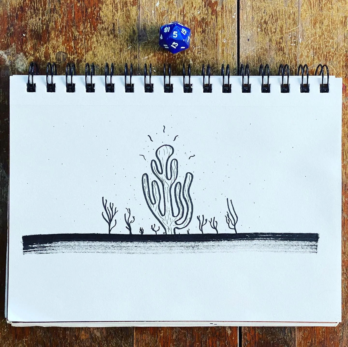 A black line drawing of stylized coral on a horizontal line is drawn in an open sketchbook placed on a rustic wooden surface, next to a blue die.