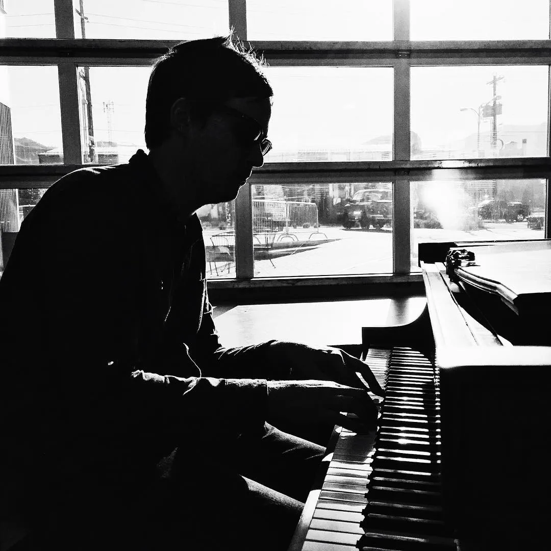 A high-contrast black and white photograph shows a silhouetted figure seated at a piano, framed by large windows overlooking a bright, urban cityscape.