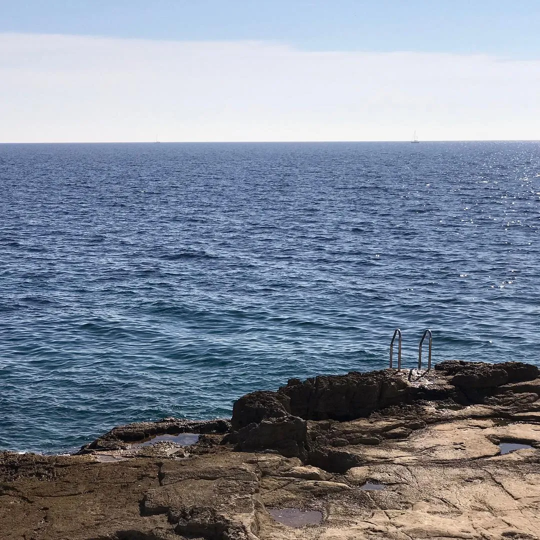 Dark, rough rocks fill the foreground, meeting a deep blue, rippling ocean that stretches toward a pale blue sky and a distant horizon.