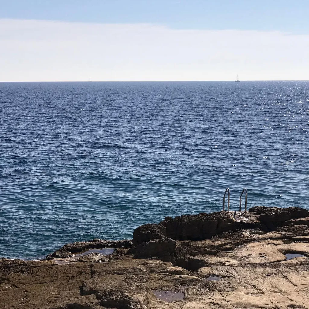 Dark, rough rocks fill the foreground, meeting a deep blue, rippling ocean that stretches toward a pale blue sky and a distant horizon.
