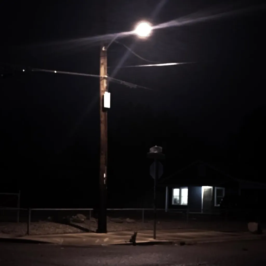 A dark night street scene dominated by a tall utility pole and a bright, glaring street lamp casting intense white light over the asphalt and a small house in the background.