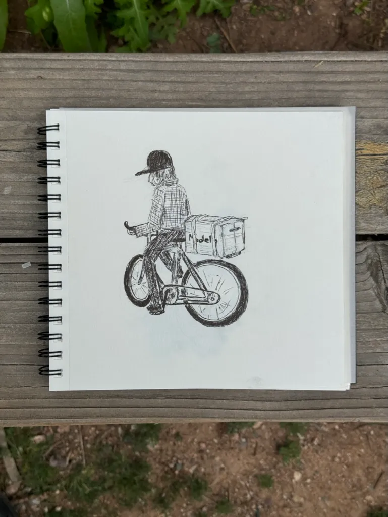 A black and gray sketch shows a figure wearing a hat riding a bicycle with a box attached, drawn on a page in a spiral-bound sketchbook resting on wood.